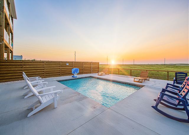 A pool with chairs and sun setting in the background.